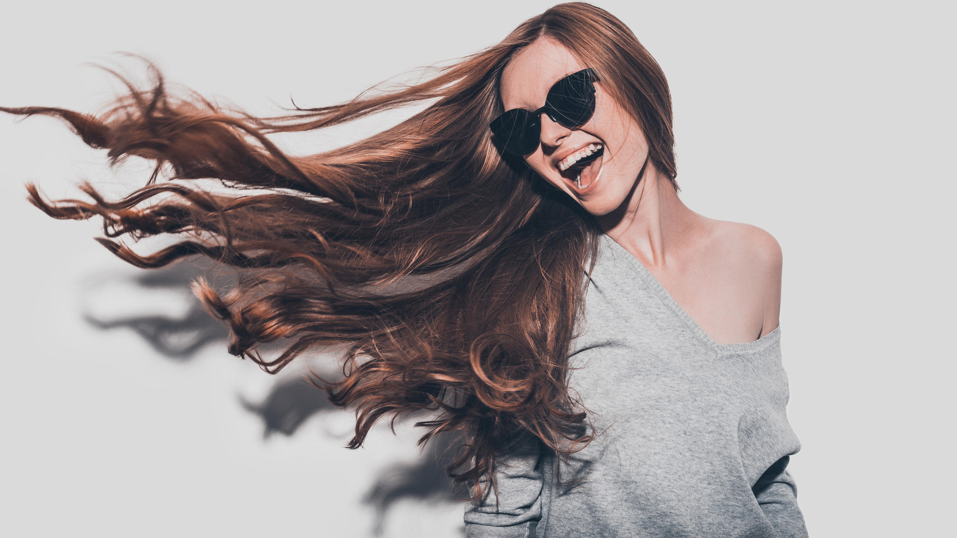 5 Haar-Hacks, die Sie kennen sollten! Attractive young smiling woman in sunglasses and with tousled hair looking away while standing against grey background
