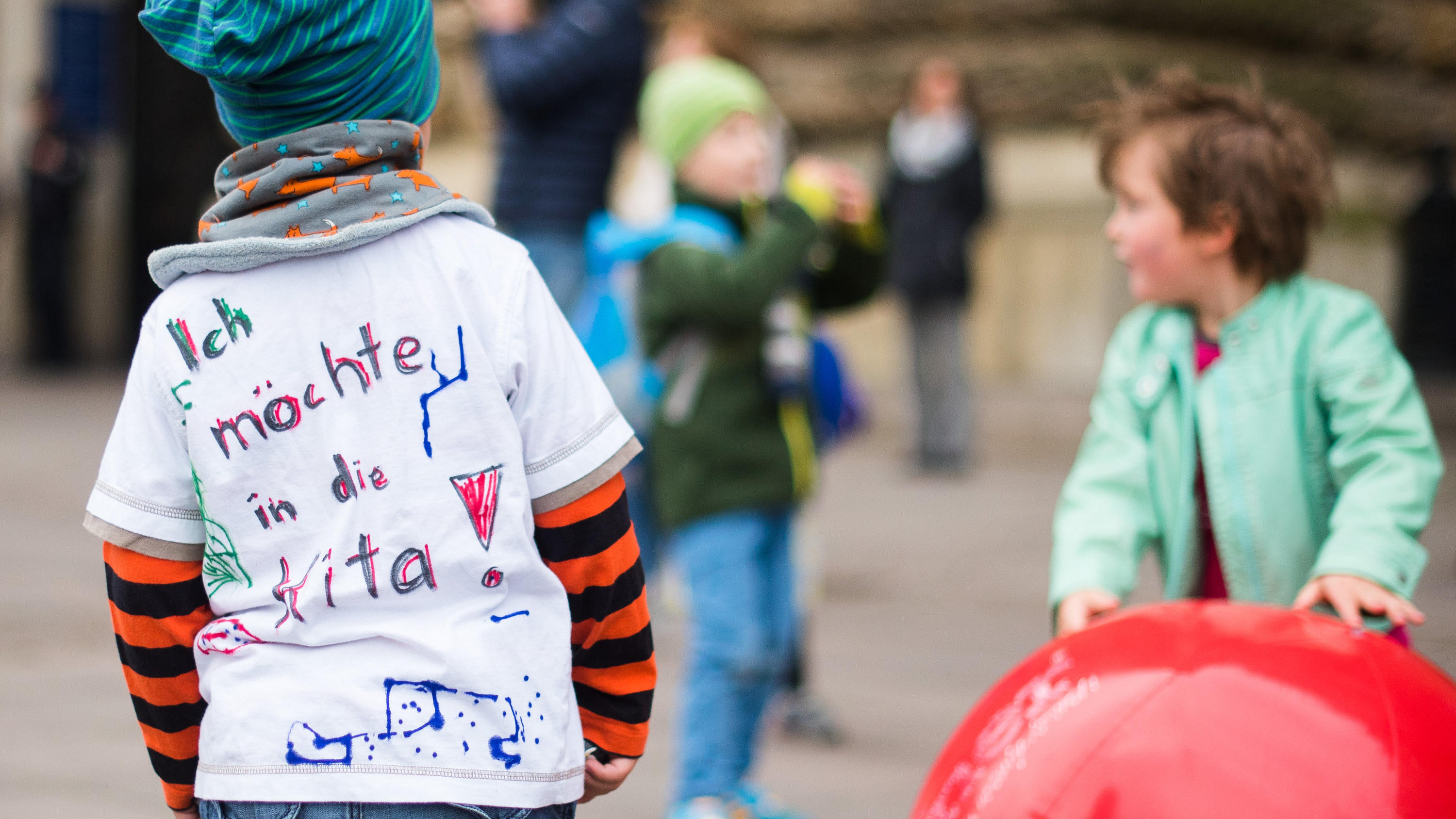 Kita-Streik: Welche Rechte haben berufstätige Eltern? ARCHIV - Ein Kind mit einem T-Shirt mit der Aufschrift «Ich möchte in die Kita» steht am 28.05.2015 in Hamburg während einer Aktion von Eltern zum Ende des Kita-Streiks vor dem Rathaus neben Kindern, die mit Bällen spielen. Foto: Daniel Bockwoldt/dpa