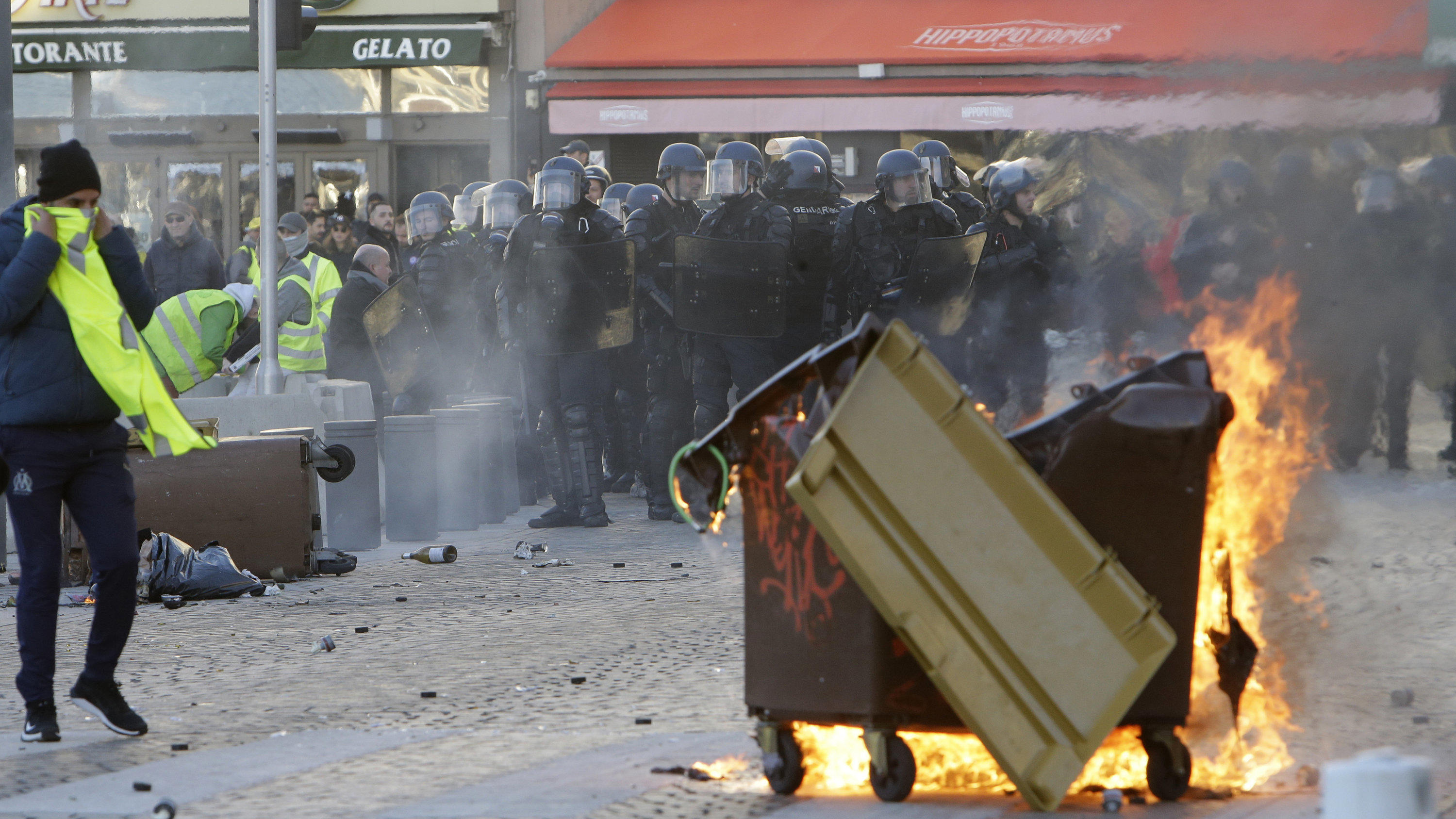 08.12.2018, Frankreich, Marseille: Die Polizei steht hinter einer brennenden Mülltonne. Bei den Demonstrationen der «Gelben Westen» in Paris hat sich die Lage am Samstagnachmittag zugespitzt. Die Polizei setzte Tränengas und Wasserwerfer ein. Nach An