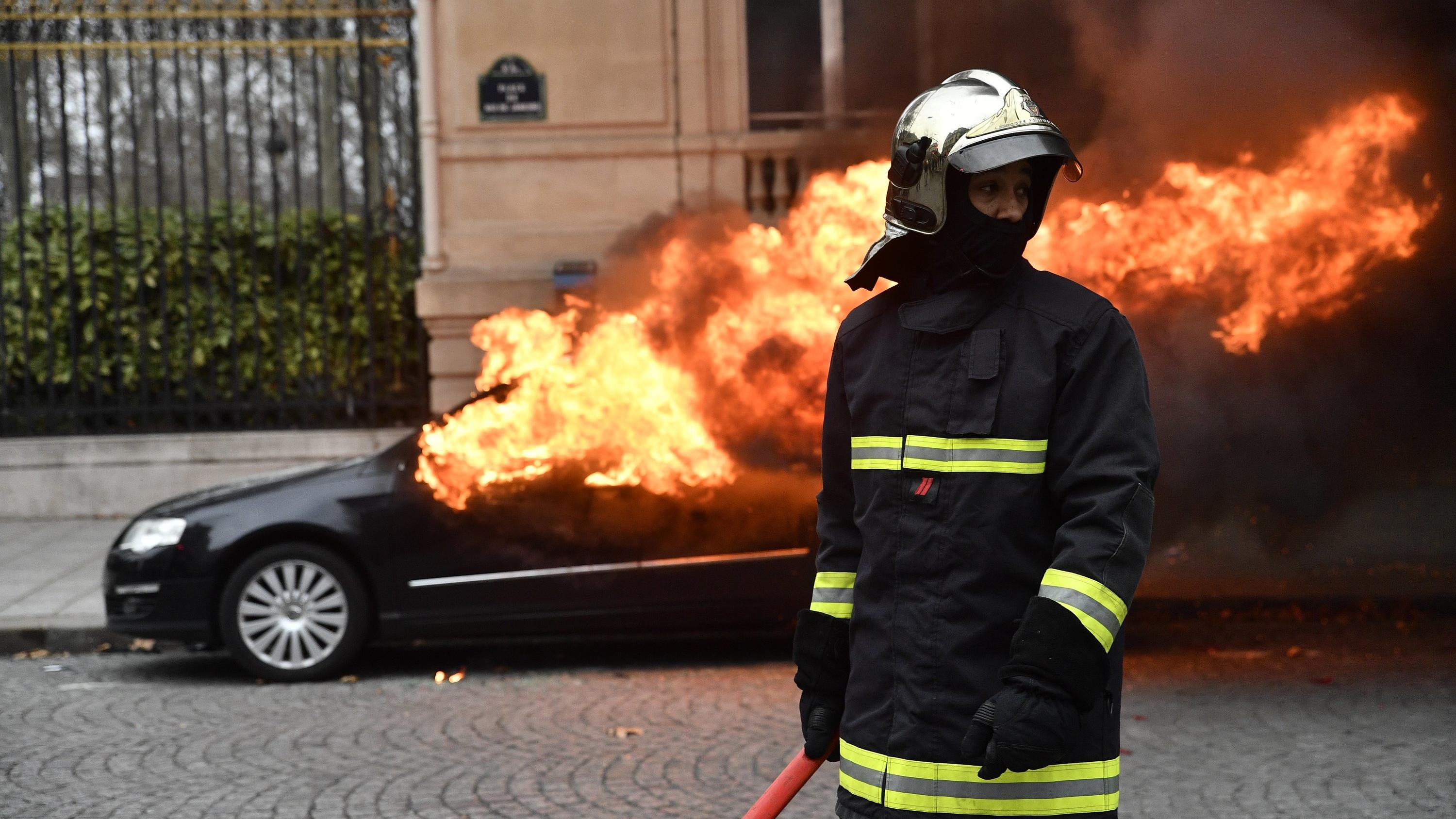 08.12.2018, Frankreich, Paris 8th (8th Arrondissement Of: Ein Mitglied der Feuerwehr steht während der Proteste und Unruhen in Paris vor einem brennenden Auto. Bei Protesten der «Gelbwesten» sind am Samstagvormittag in Paris mehrere Hundert Menschen